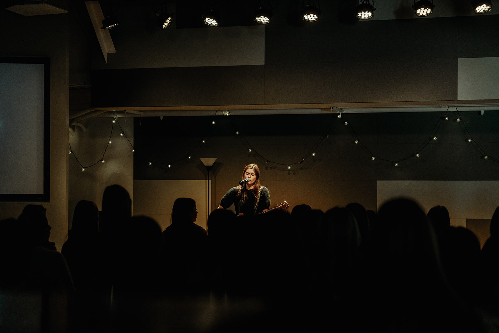oung woman singing and playing guitar on stage under soft lights as students gather for a High School Ministry worship service.