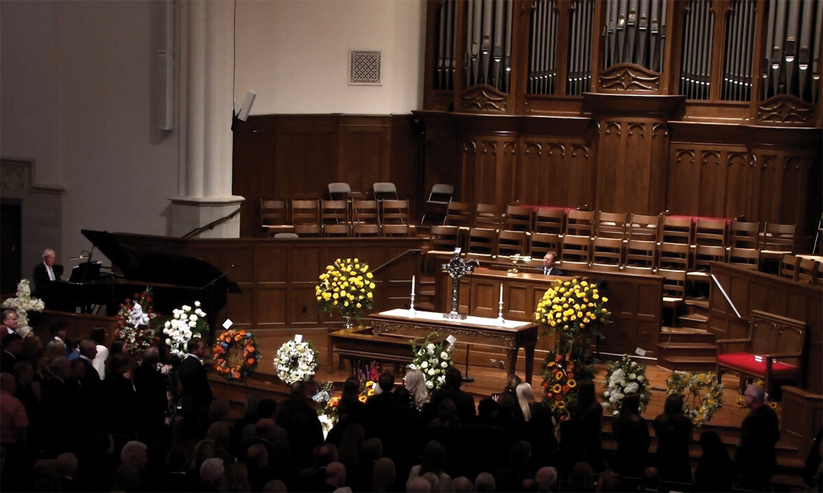 Congregation gathered in a church sanctuary for a memorial service, with a casket at the front surrounded by floral arrangements.