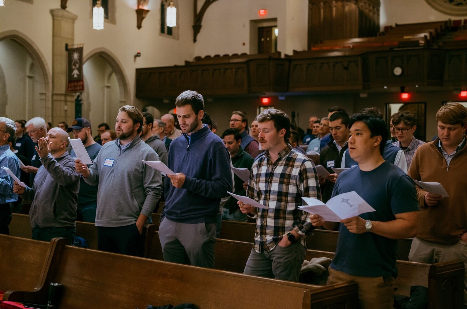 Men standing in church pews singing from printed programs during a worship service.