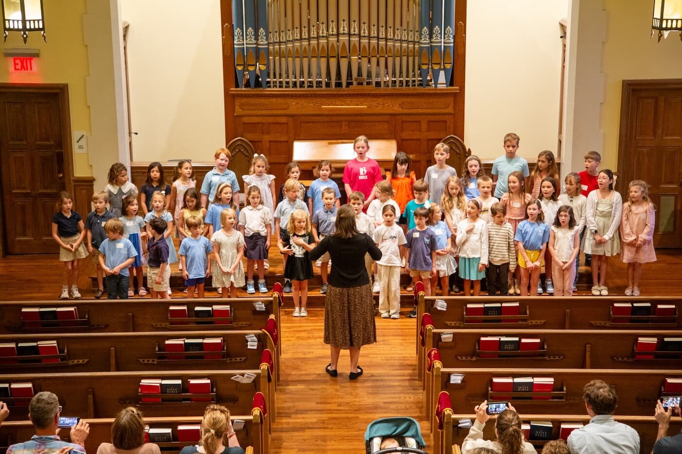 Children’s choir performing in a church sanctuary, directed by a music leader in front of the congregation, with a pipe organ visible behind them.