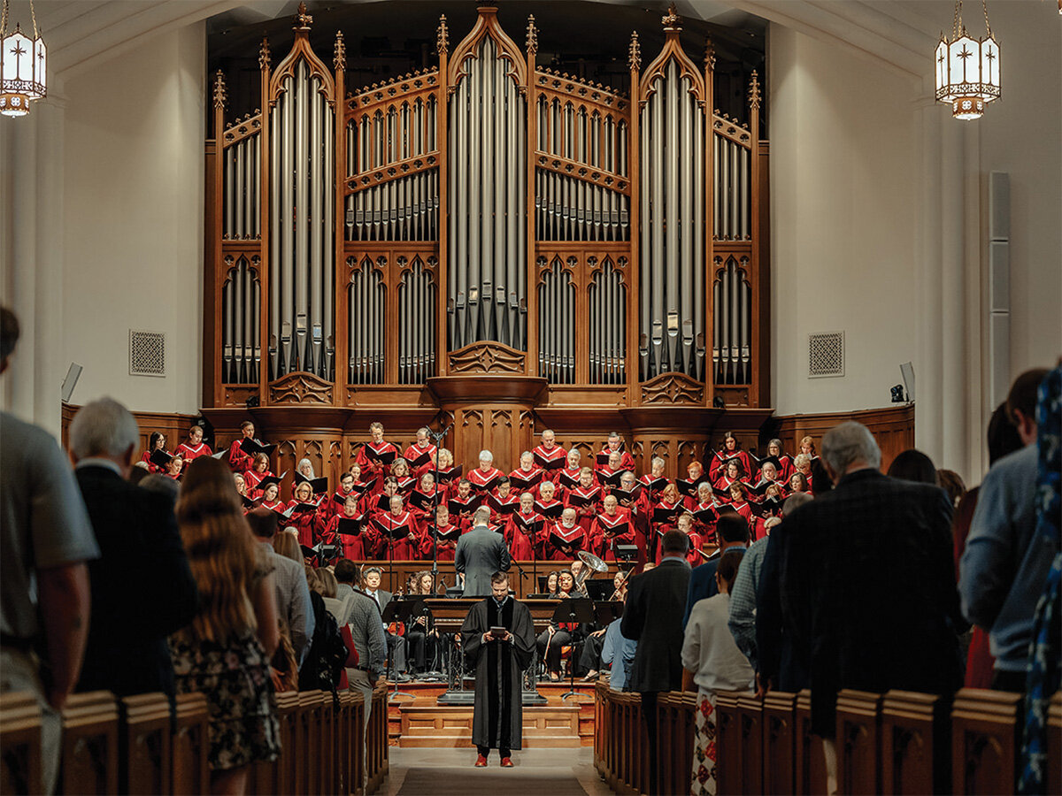 Church choir in red robes singing in the sanctuary beneath a large pipe organ, with the congregation standing during worship.