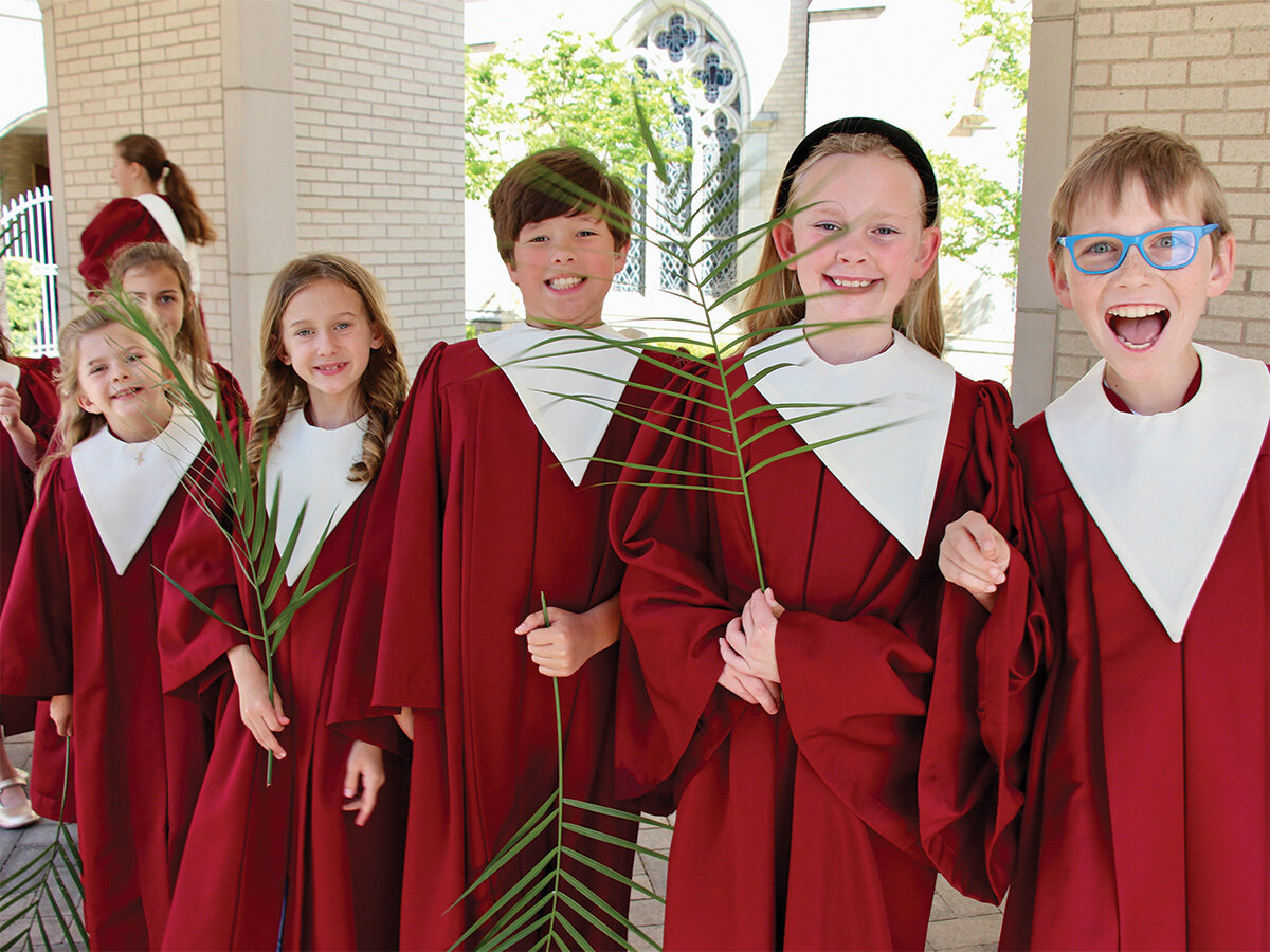 Children in red choir robes holding palm branches outside a church, smiling during a Palm Sunday celebration.
