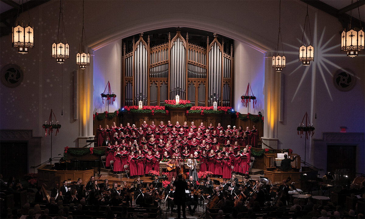 Large church choir in red robes singing in front of a pipe organ, accompanied by an orchestra.